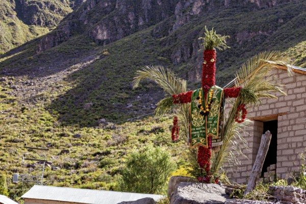 A scarecrow decorated with traditional costumes stands in a sunny mountain landscape next to a small building, cross in the landscape of the Colca Canyon of Peru