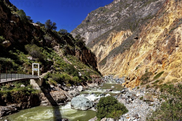 Narrow bridge across river flanked by steep rock walls under deep blue sky, the landscape of Colca Canyon in the Aden of Peru