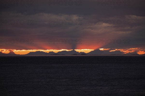 The Vestfjord in Nordland, Norway, offers a dramatic atmosphere near Bodø. Dark clouds are illuminated by the setting sun in shades of red and yellow. The rugged cliffs and mountains are reflected in the quiet fjord, which offers a breathtaking view of Lofoten