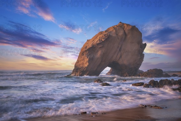Penedo do Guincho, a large boulder rock arch at Praia da Santa Cruz, Portugal, with ocean waves and sandy beach on sunset