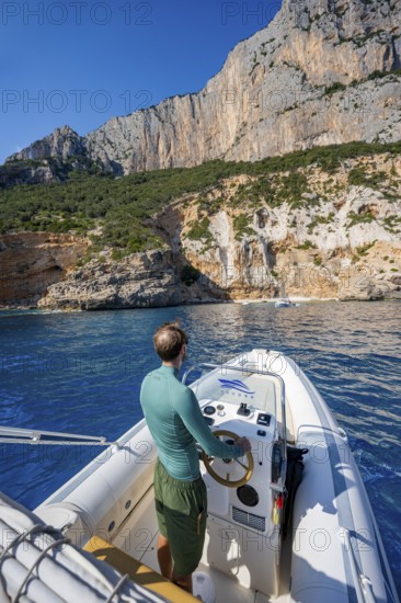 Young man rides a motorboat along the picturesque rocky coast, cliffs and blue sea, Golfo di Orosei, Baunei, Sardinia, Italy