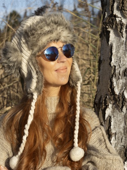 Slender pretty woman with fur hat and pompoms, red hair and warm winter colors between birch forest and reeds