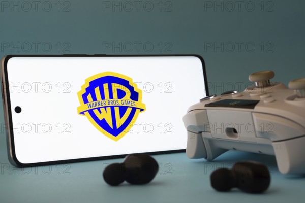 Dhaka, Bangladesh- 23 Mar 2025: A smartphone with a Warner Bros. Games icon next to a controller and earbuds on a blue surface