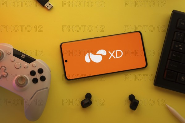 Dhaka, Bangladesh- 22 Mar 2025: Smartphone with XD logo, game controller, and earbuds on a pink background