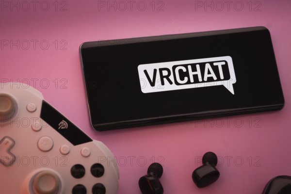 Dhaka, Bangladesh- 22 Mar 2025: Smartphone with VRChat logo, game controller, and earbuds on a pink background