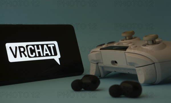 Dhaka, Bangladesh- 23 Mar 2025: A smartphone with a VRChat icon next to a controller and earbuds on a blue surface
