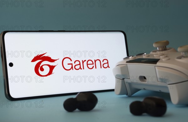 Dhaka, Bangladesh- 23 Mar 2025: A smartphone with a Garena icon next to a controller and earbuds on a blue surface