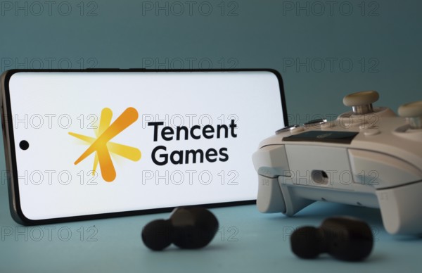 Dhaka, Bangladesh- 23 Mar 2025: A smartphone with a Tencent Games icon next to a controller and earbuds on a blue surface