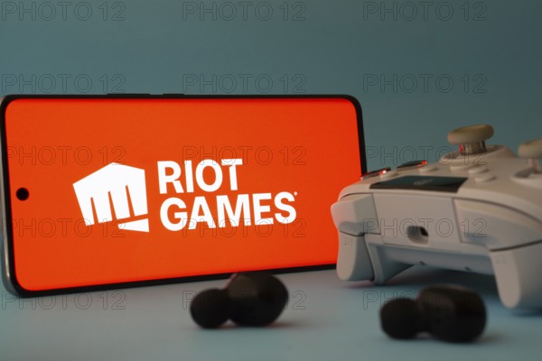Dhaka, Bangladesh- 23 Mar 2025: A smartphone with a Riot games icon next to a controller and earbuds on a blue surface