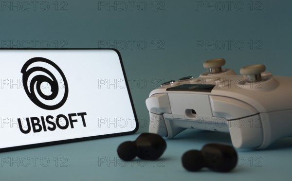 Dhaka, Bangladesh- 23 Mar 2025: A smartphone with a Ubisoft icon next to a controller and earbuds on a blue surface