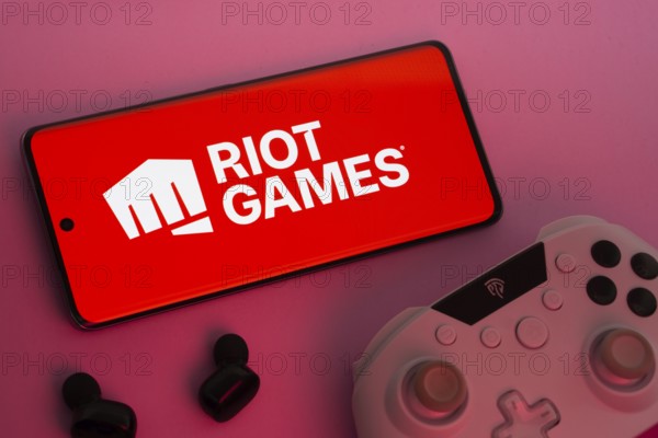 Dhaka, Bangladesh- 22 Mar 2025: Smartphone with Riot Games logo, game controller, and earbuds on a pink background