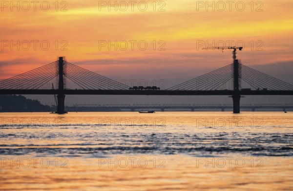 A boat sails on the river Brahmaputra during sunset, with the silhouette of a cable-stayed bridge and hills in the background, on November 4, 2025 in Guwahati, India