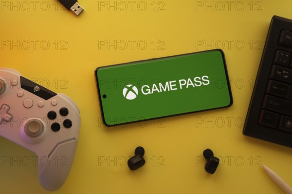 Dhaka, Bangladesh- 15 July 2025: Smartphone with Xbox game pass logo, game controller, and earbuds on a yellow background
