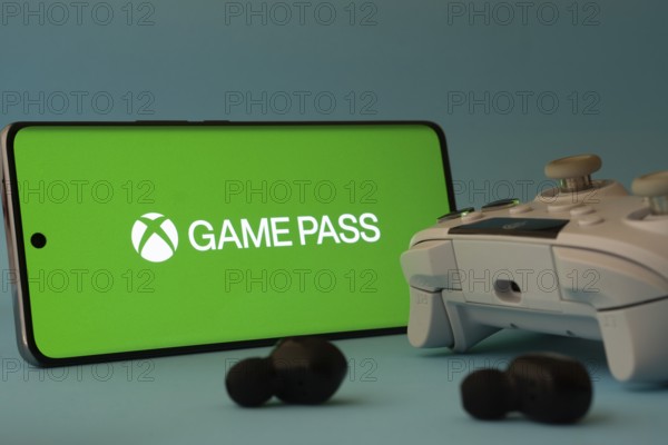 Dhaka, Bangladesh- 15 July 2025: A smartphone with a Xbox Game Pass icon next to a controller and earbuds on a blue surface