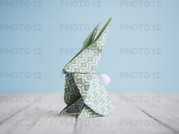 Green origami bunny standing on a white wooden table with a light blue background, representing easter and creativity