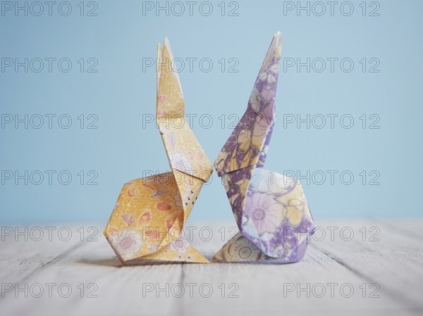 Two colorful origami bunnies are facing each other, touching noses, on a wooden surface with a light blue background