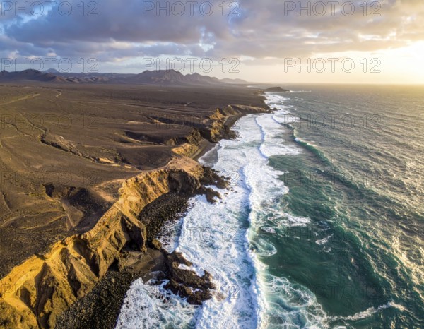 Desert landscape converges with ocean along a rugged coastal area, shore break at cliffs, blue cloudy sky at sunset, dry landscape in an aerial drone view, AI generated
