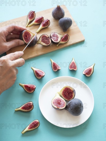 Whole and sliced fresh figs on a blue background, with hands cutting them on a wooden board