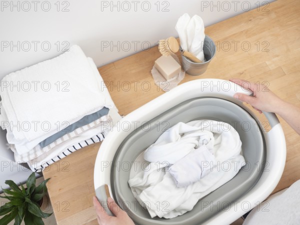 Housekeeper holding a basin with white clothes, soap bar and brush on wooden table, preparing to wash laundry