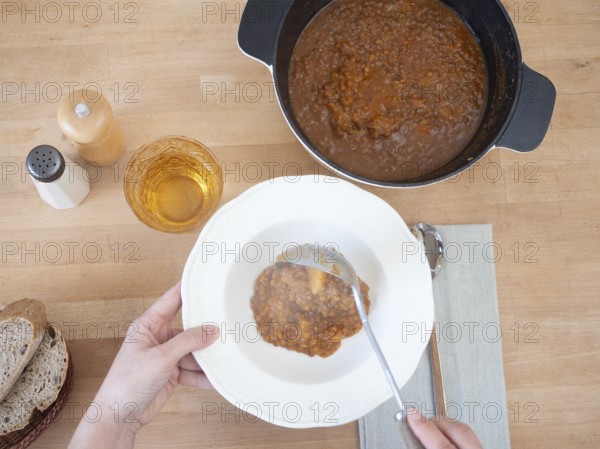 Hands serving lentil stew with ladle from pot to bowl, with bread, water and salt and pepper shakers on wooden table