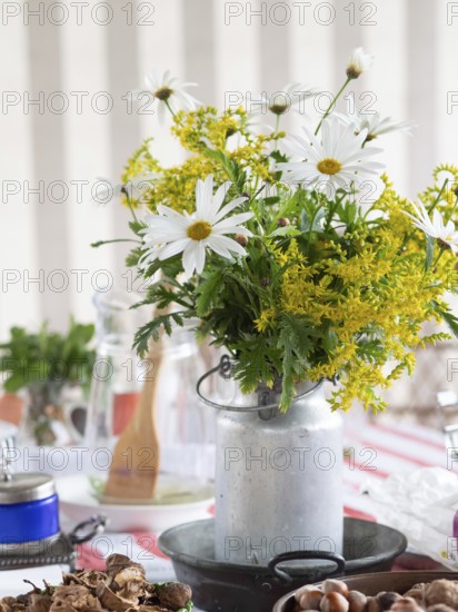 White daisies and yellow goldenrod create a vibrant centerpiece in a rustic milk can, adding a touch of nature to a table setting adorned with nuts and kitchen utensils