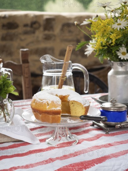 Slice of lemon cake with powdered sugar on a glass stand, pitcher of lemonade, flowers, and colorful tablecloth