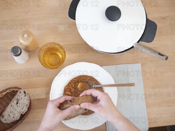 Hands breaking slice of bread over bowl of lentil soup, served with drink, salt and pepper, and bread basket, top view