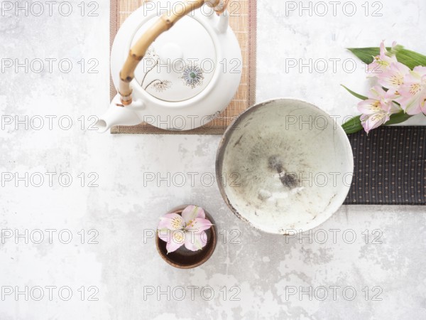 Asian tea ceremony arrangement with white teapot, empty bowl, pink flowers and bamboo placemat on white background
