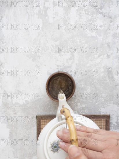 Hand gracefully pours tea from a white ceramic teapot with bamboo handle into a small brown bowl on a textured gray surface