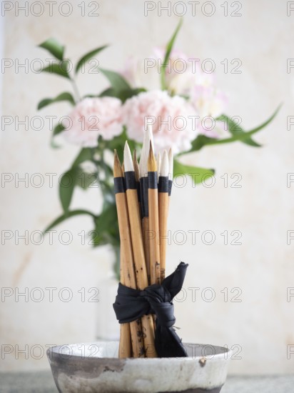Calligraphy brushes are tied with a black ribbon and placed in a ceramic bowl, with pink carnations and alstroemeria flowers softly blurred in the background