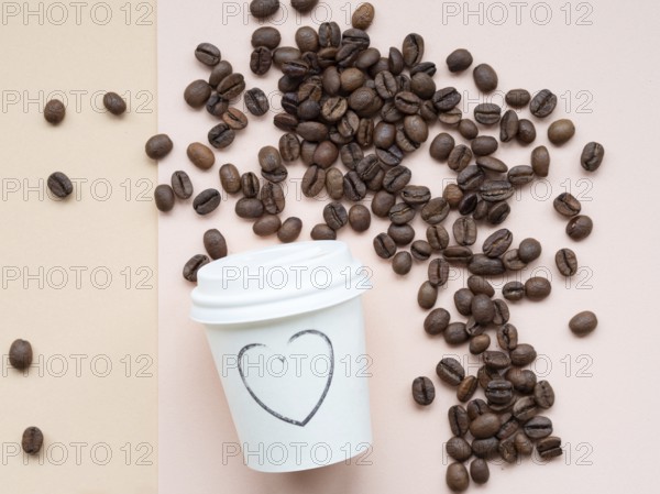 Roasted coffee beans scattered on a two-tone background near a paper cup with a heart drawn on it, conveying a love for coffee