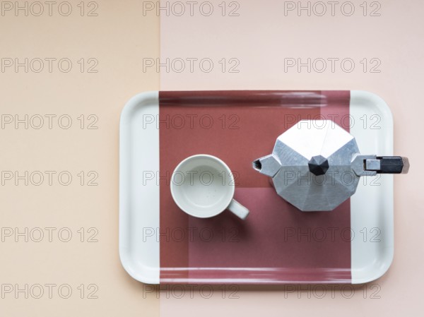 Overhead view of a moka pot and an empty coffee cup sitting on a serving tray, arranged on a two-toned background, creating a minimalist and inviting scene