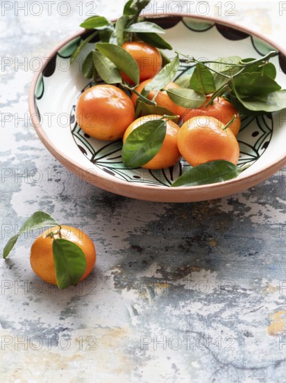 Vibrant tangerines with attached leaves are nestled in a ceramic bowl, placed on a textured, rustic surface, creating a simple yet appealing still life