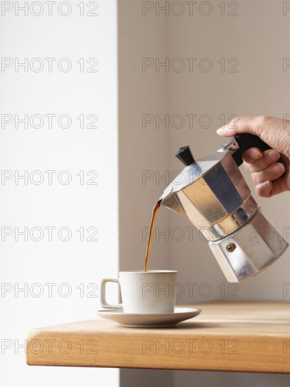 Hand pouring coffee from a moka pot into a cup, placed on a wooden table, creating a cozy and aromatic scene