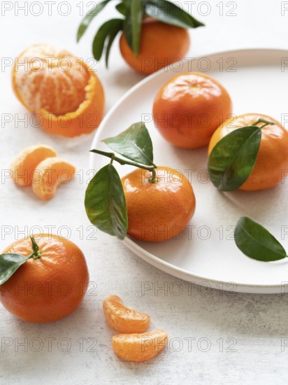Composition of tangerines with leaves, some peeled and segmented, arranged on a white plate and table
