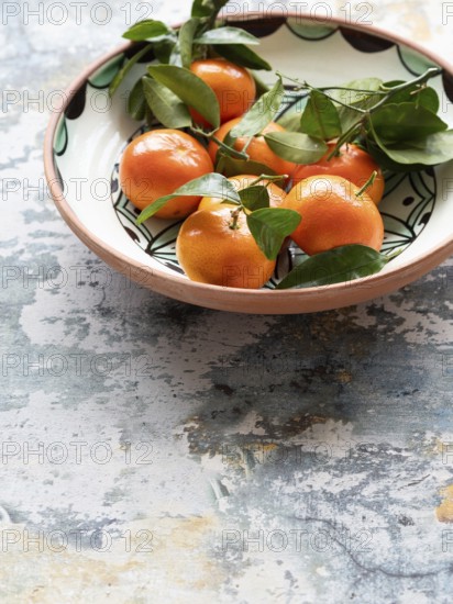 Vibrant tangerines with green leaves arranged in a decorative ceramic bowl, set against a rustic tabletop, creating a still life composition