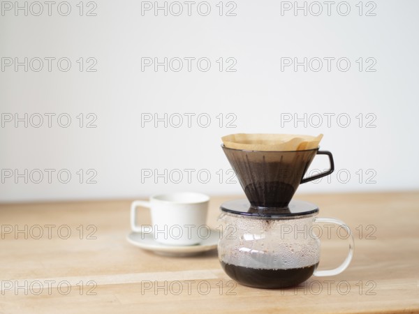 Coffee is brewing using a paper filter and dripper on a wooden table, with a white cup and saucer nearby