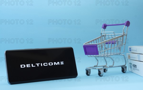 Dhaka, Bangladesh- 03 Oct 2025: Smartphone with Delticom logo next to a small shopping cart and boxes against a blue background