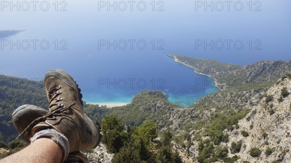 A person relaxes while hiking on a steep cliff in the mountains with spectacular views of hiking boots and the sea, the coastline and the blue water, Turkish Aegean Sea, hiking the Lycian Way, Turkey