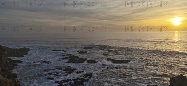 The picture shows a sunset over the sea with rocks in the foreground, hiking on the Fisherman's trail, Rota Vicentina, Porto Covo, Alentejo region, Portugal