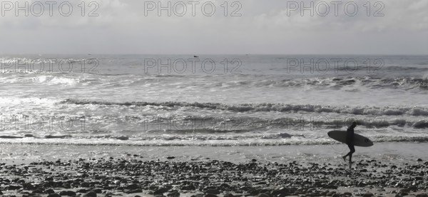 Silhouette of a surfer carrying his board along a pebble beach with a view of the wavy ocean, Sidi Ifni, Morocco
