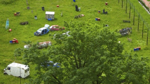 Green field with scattered car wrecks and barrels surrounded by trees, Poland