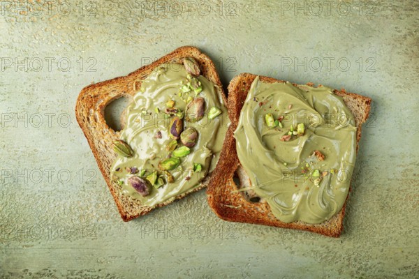 Two slices of toasted bread, topped with pistachio paste and sprinkled with pistachios, spread on a light surface