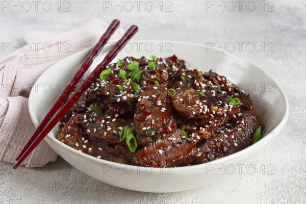Crispy beef, with green onions and sesame seeds, in a bowl with chopsticks, on a textured surface, on a light background, no people