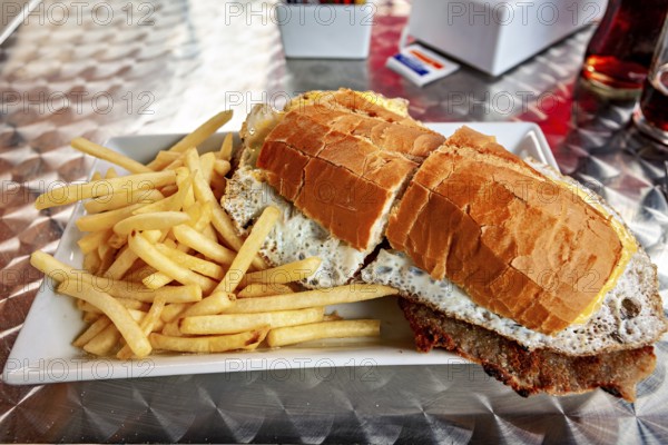 Large sandwich with fries served on a metal table, thickly filled with bread and a fried egg visible, An Argentinean Lomito
