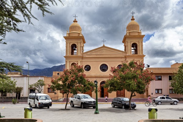 Church façade with two bell towers surrounded by trees and cars, under a dramatic sky, the Catedral de Nuestra Señora del Rosario in Cafayate, Argentina