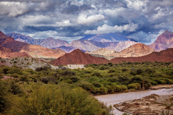 River flows through green vegetation against a mountain backdrop with red hills, the landscape and colorful rock formations of the Quebrada de Cafayate near Salta in Argentina