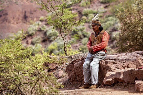 A traditionally dressed man sits thoughtfully on a rock in a desert landscape, a local in the rock formation of the Quebrada de Cafayate near Salta in Argentina