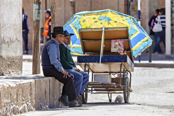 Two men resting under a blue parasol next to a cart, people in the streets of San Antonio de los Cobres, Salta province, Argentina