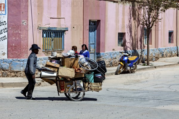 Man pulls carts full of packages past a colorful building, people in the streets of San Antonio de los Cobres, Salta province, Argentina